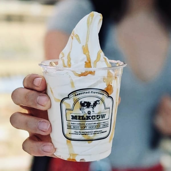 Hand holding a clear cup of soft-serve ice cream drizzled with honey, with a Milkcow logo.