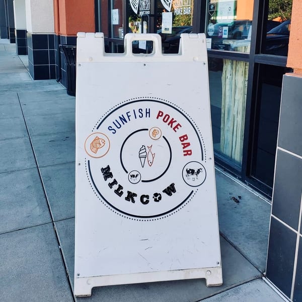 A white A-frame sign for Sunfish Poke Bar and Milkcow stands on a sidewalk.