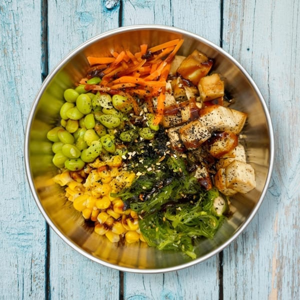 A Sunfish Poke Bar bowl with edamame, corn, seaweed salad, carrots, and tofu, on a blue wooden table.