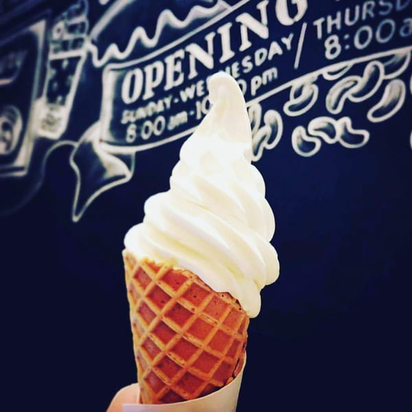 A waffle cone with soft-serve ice cream held against a dark background with white chalk writing.