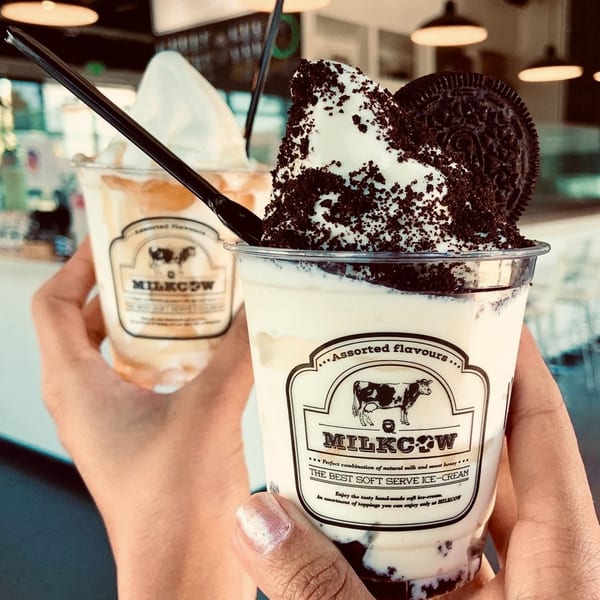 Two hands holding Milkcow soft-serve ice cream cups, one with Oreo crumbles and a cookie.